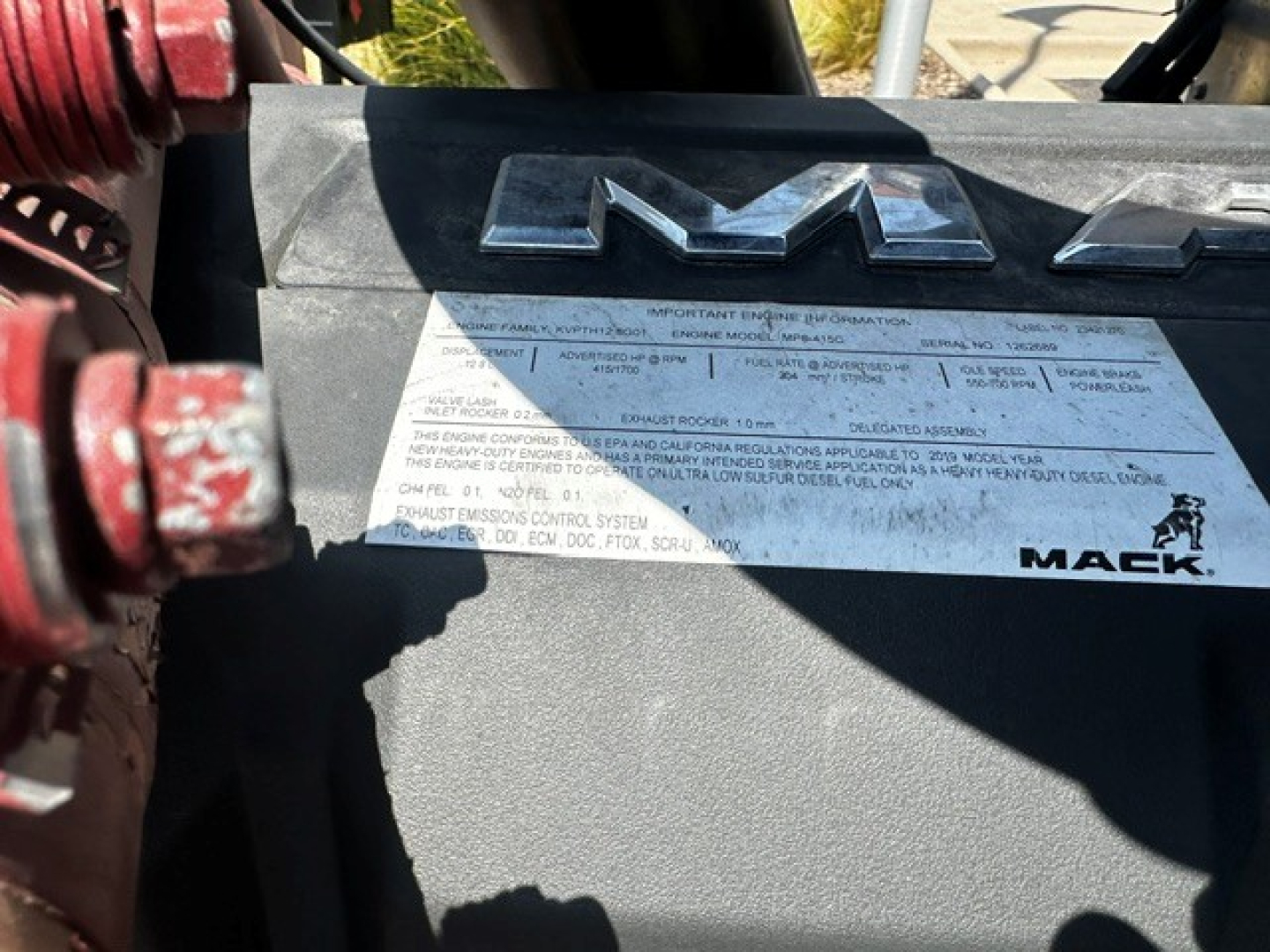 Truck engine bay showing a large metallic Mack logo on a label with small text and VIN plate.