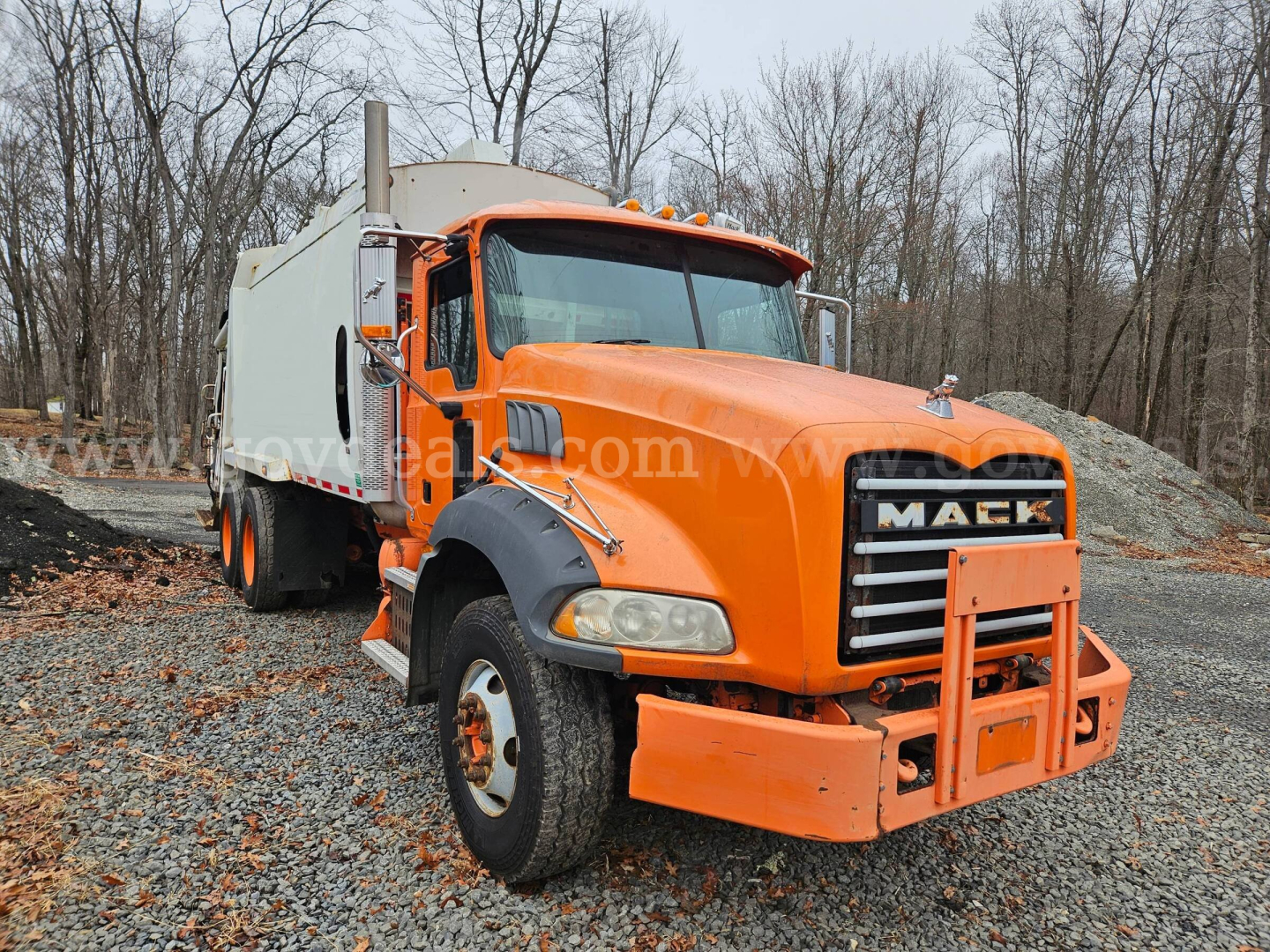 Mack GU813 truck in an outdoor lot, orange cab with large front bumper guard and gray waste body; visible VIN tag and stock number marking in image.