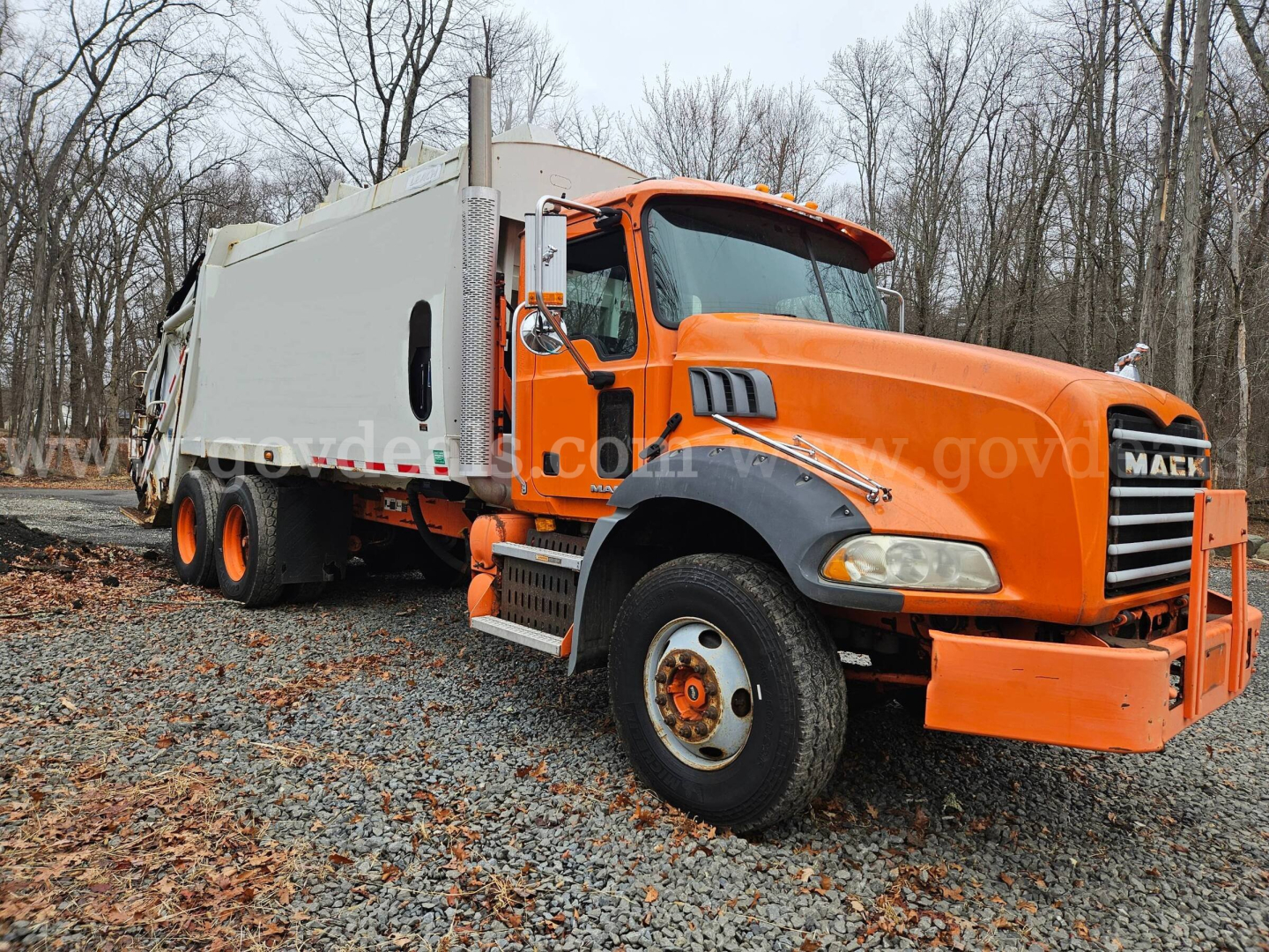 Orange Mack GU813 refuse truck with white rear body, front view showing cab and large bumper, VIN 1M2AX13C79M008139 visible, on gravel with leaf litter.