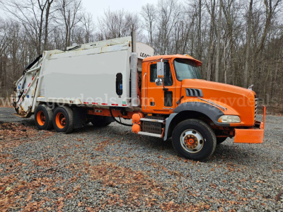 Mack GU813 garbage truck in-service showing orange cab and white rear body with three-axle configuration on gravel lot.