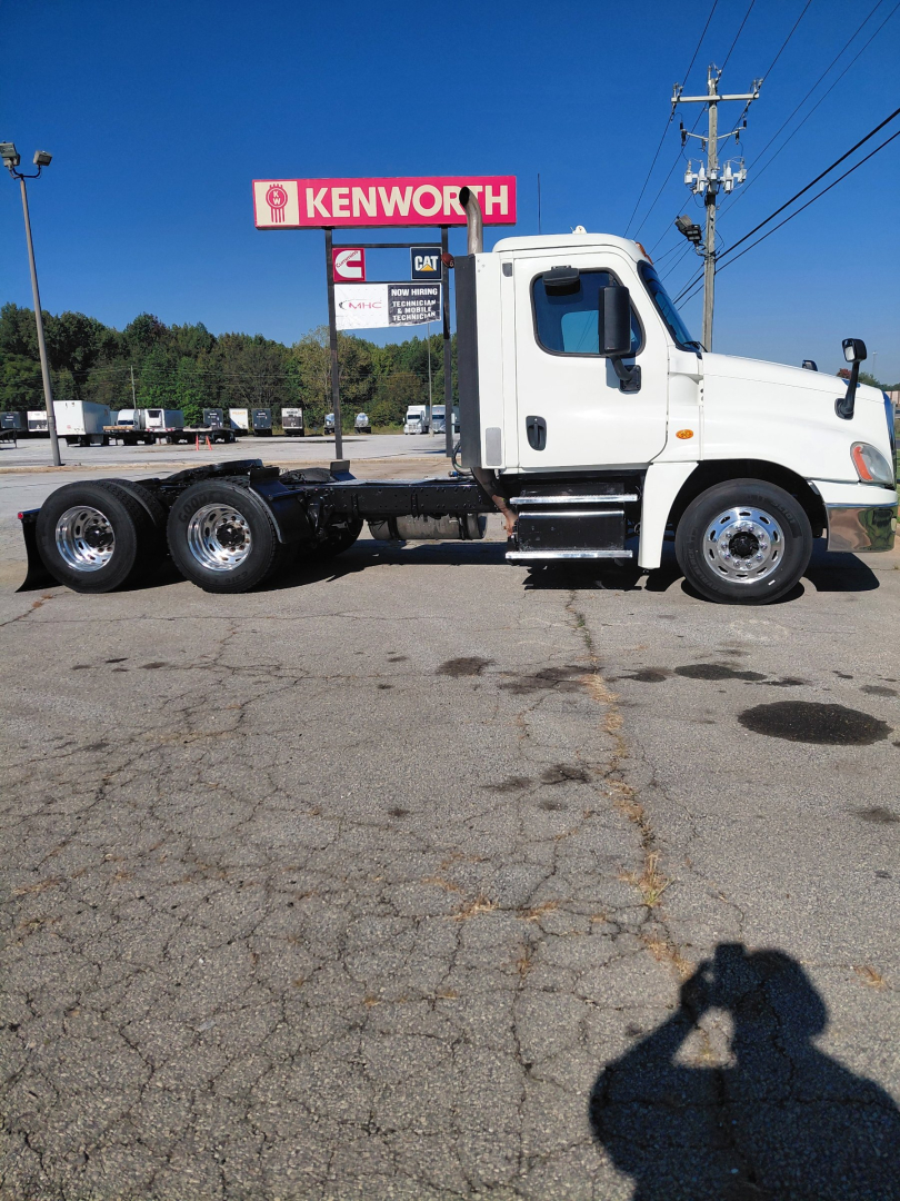 Truck chassis view of a Freightliner PT126064S T conventional sleeper with three rear axles and white cab, showing fuel tanks, step, and Kenworth sign in background.