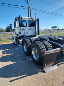 Truck rear-angle view of a white Freightliner PT126064S tractor showing dual rear axles with chrome wheels and a black mud flap with MHC logo.