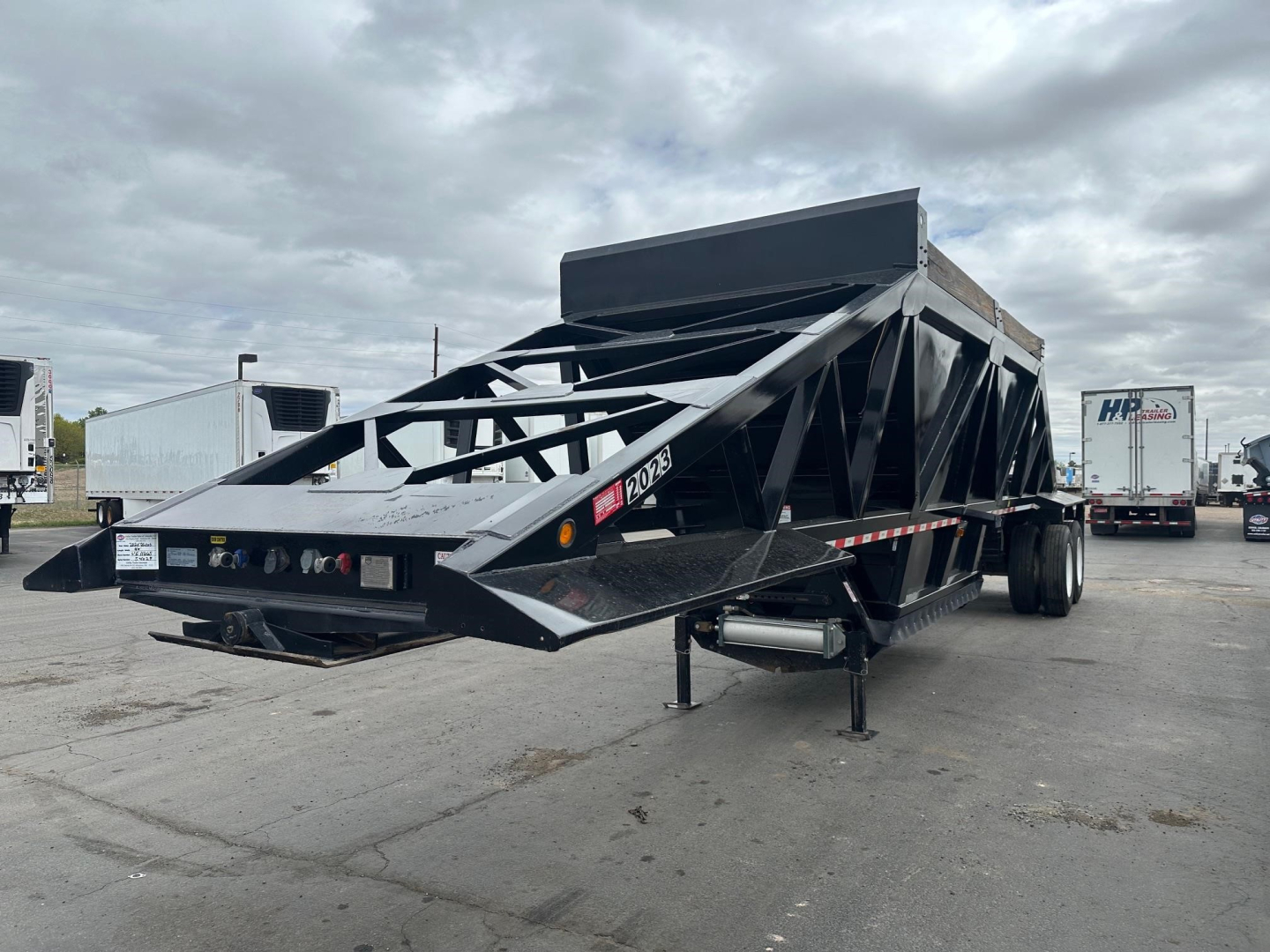 Trailer shows bottom dump semi-trailer with black steel frame and raised side walls; visible labels and lights on rear; loading area and support legs in foreground.
