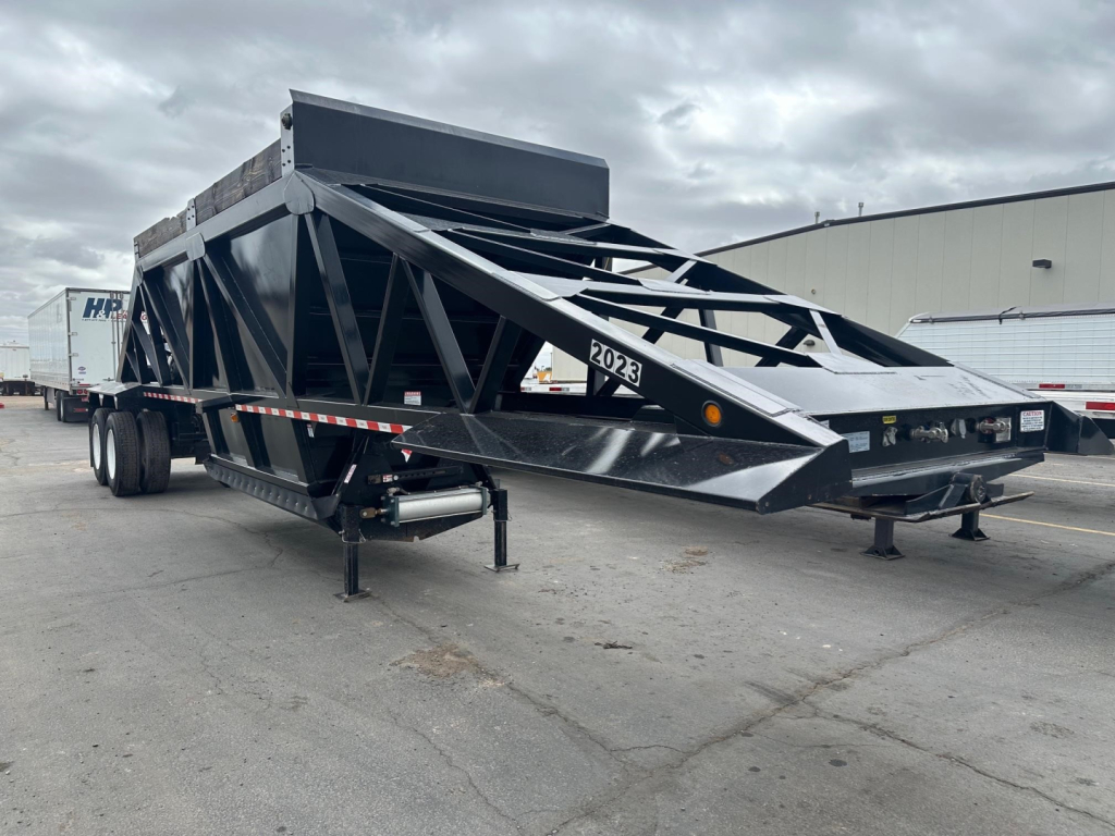 Brazos bottom dump trailer in view showing structural lattice frame, dual axle, and front hopper. Visible text includes '2023' on the side panel and a sticker with the number.