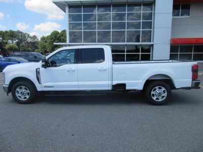 Side view of a white Ford F-250 pickup truck in a dealership lot, long crew cab with four doors and long cargo bed, dealership building in background.
