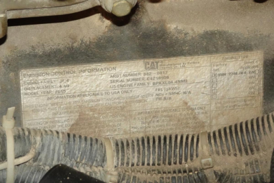 Emissions control information plate on equipment showing CAT serial and engine numbers with model and date codes.