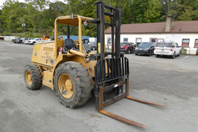Forklift in outdoor lot showing yellow Master Craft C06-10116 with black mast forks and tires, lifting carriage at foreground; several vehicles and a building in background.
