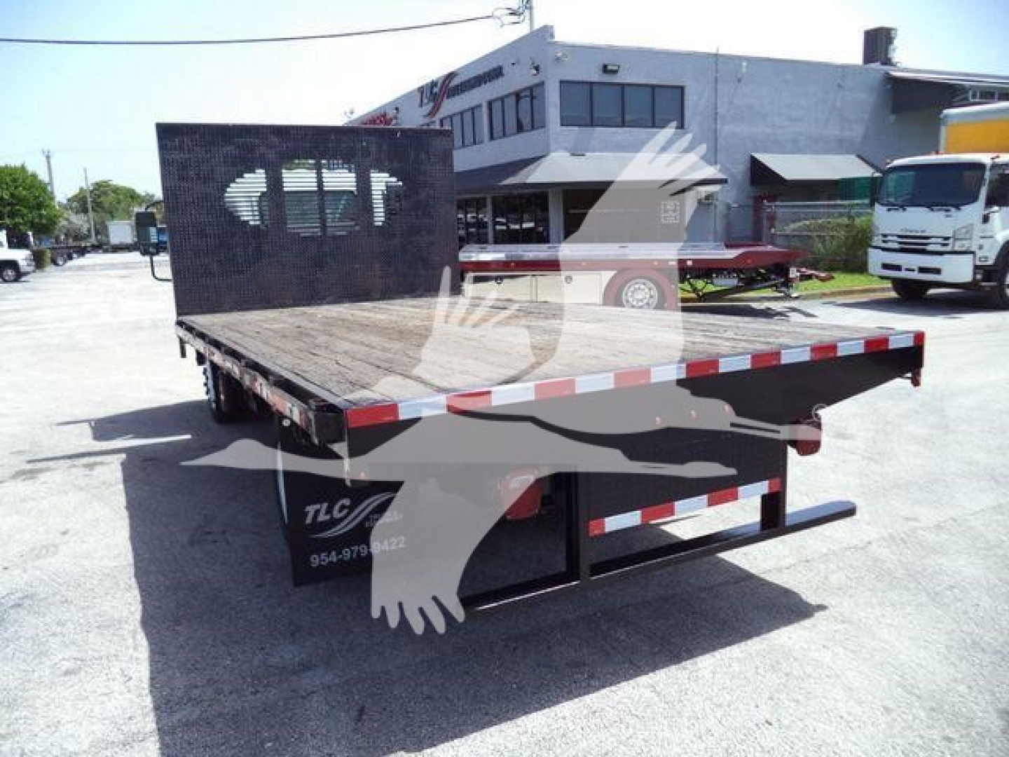 Flatbed dump truck bed on Isuzu NPR flatbed with open rear, metal deck, and black partition screen; orange and white reflective tape along edges; truck yard in background.