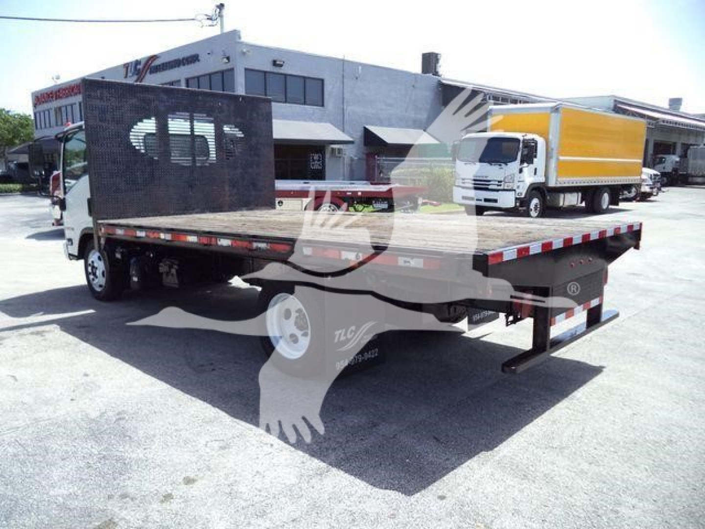 Isuzu flatbed dump truck in a dealership lot with a yellow box truck in background; flatbed has black screen divider and rear ramps.