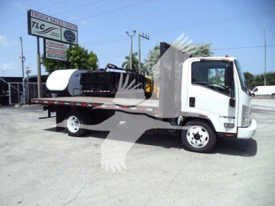 Isuzu NPR flatbed tow truck with a white cab and flatbed, showing a fuel or hydraulic tank on the bed and a sign in background. Visible text includes a TCL Truck Sales Center sign and various dealership details; license plate area not legib