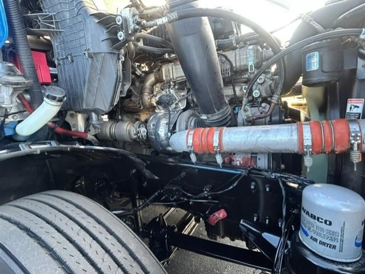Engine bay of a Freightliner Cascadia showing various hoses, engine components, and an air dryer labeled WABCO with a cylindrical canister; visible oil lines and a red pipe wrap.