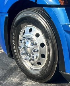 Truck wheel close-up showing large steel rim with multiple lug nuts and tire with visible tread. Rim has circular holes, and tire sidewall displays markings.