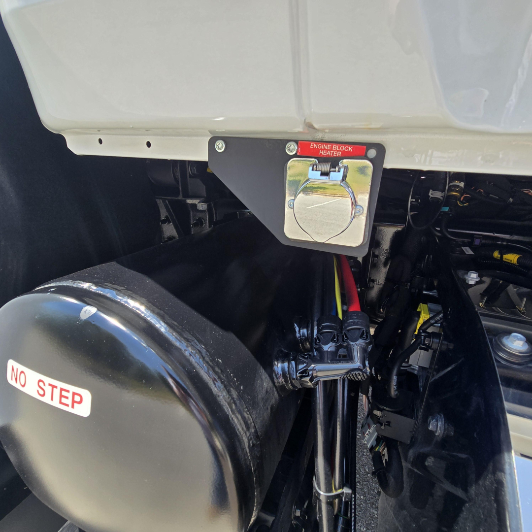 Dump truck rear showing engine block heater inlet cover, hydraulic lines, and a black cylindrical air tank with 'NO STEP' sticker.