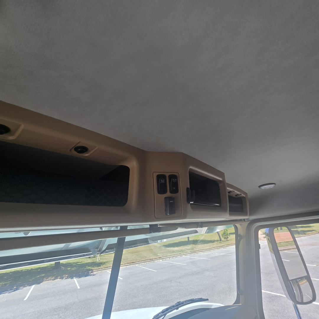 Cab interior overhead storage and controls in a Mack PI64 truck cab, showing beige panels, small switches, and a window mirror visible through the windshield.