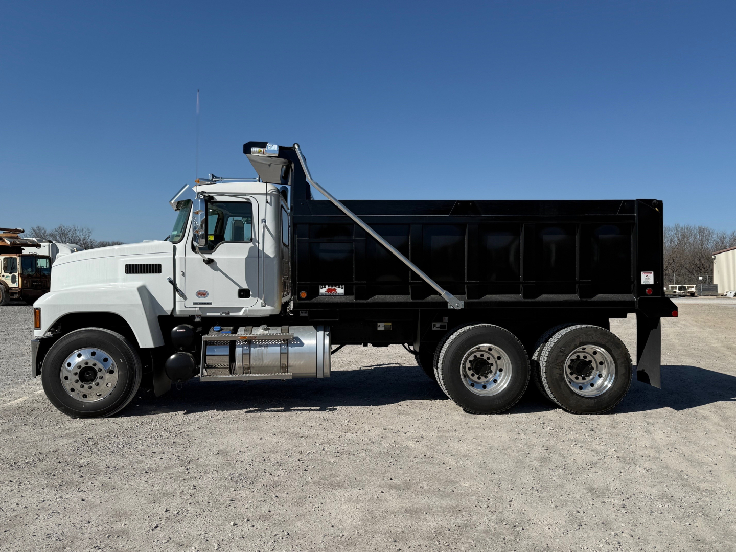 Dump truck side view showing Mack PI64 cab and black dump bed with three-axle rear. Visible text on sticker near cab and drivetrain chrome exhaust, no specific model text readable.