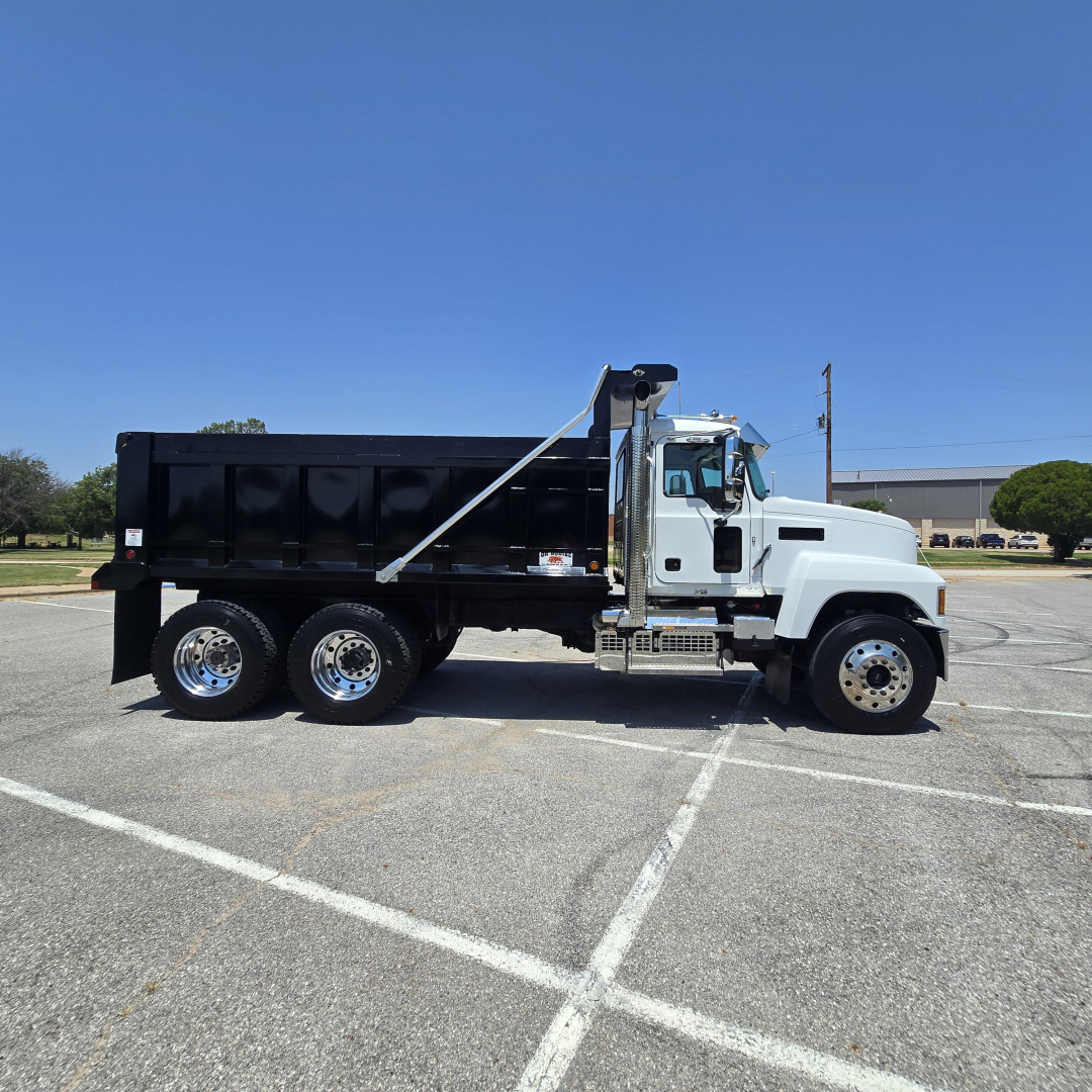 Truck side view of a white Mack PI64 dump truck with black dump bed, six wheels, and hydraulic dumping mechanism.