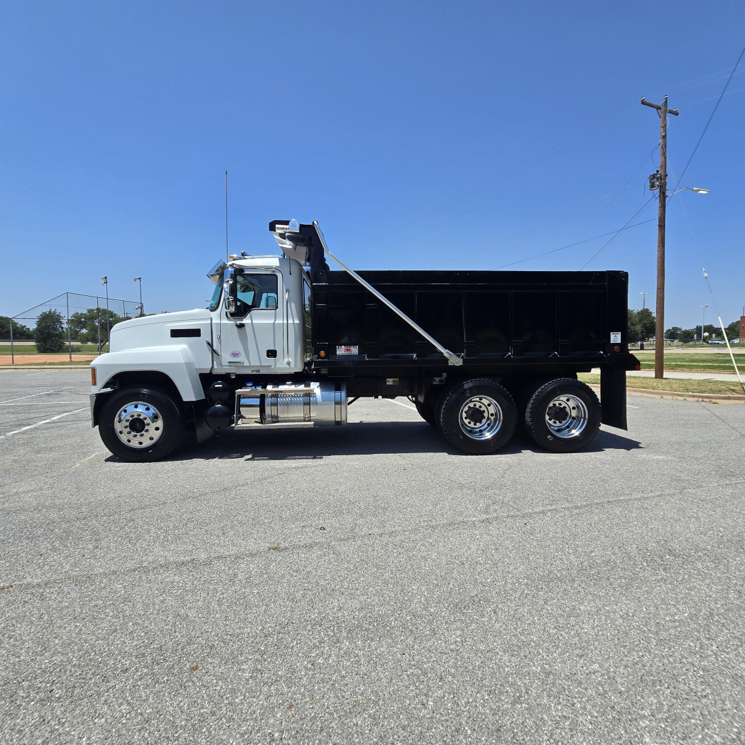 Side view of a new Mack PI64 dump truck with a black dump bed, silver cab, and tandem rear axles.