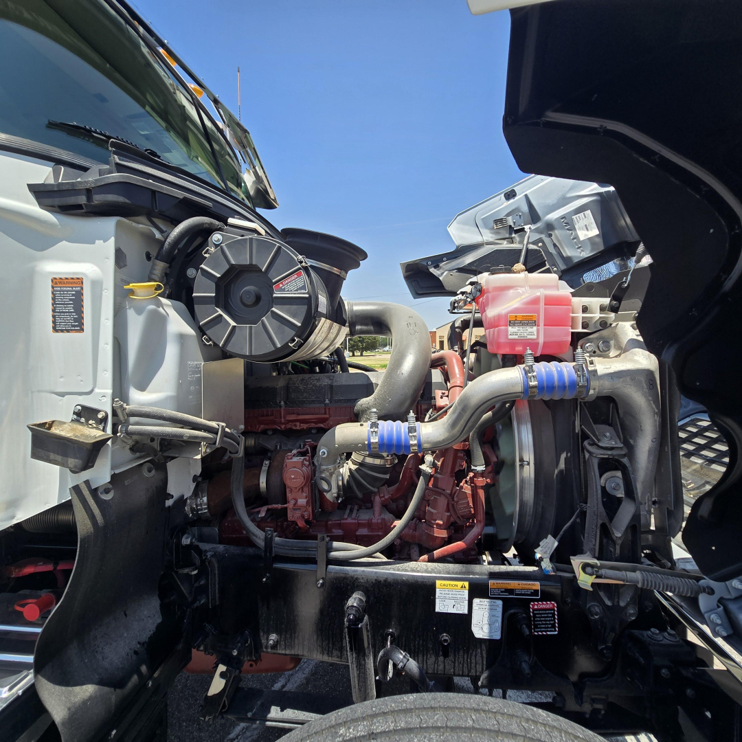 Front engine bay of a Mack PI64 dump truck showing radiator, hoses, coolant reservoir, and engine block visible.
