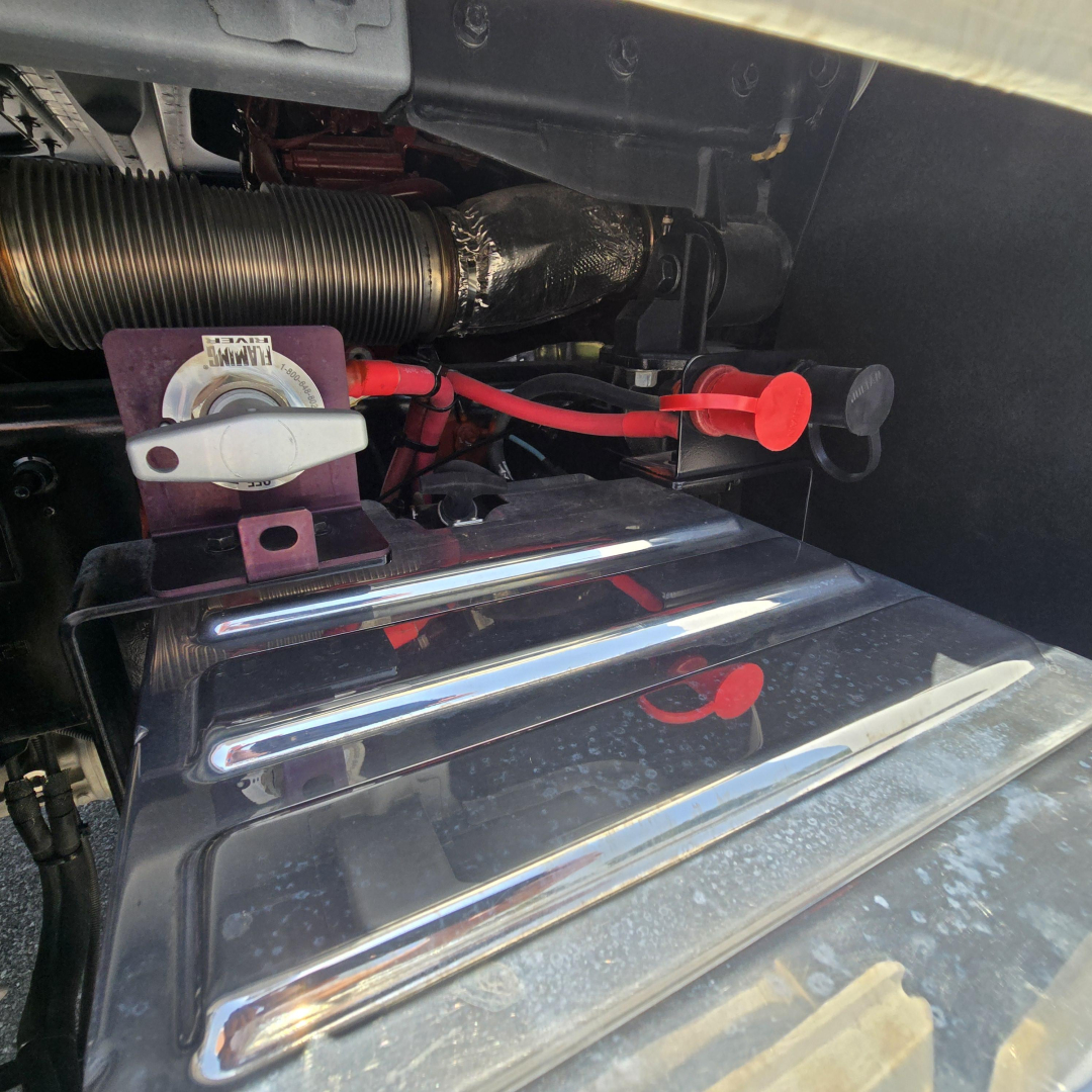 Mack PI64 engine bay shows battery quick-connect red clamps and chrome fuel tank cover with wiring and hoses in view.
