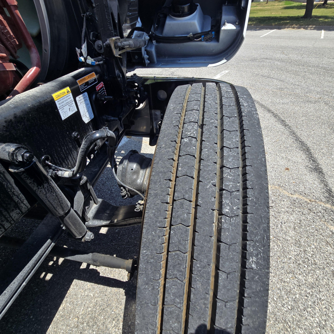 Truck front wheel and chassis showing dual tread tires and frame hardware on Mack PI64 in-service area with warning labels visible.