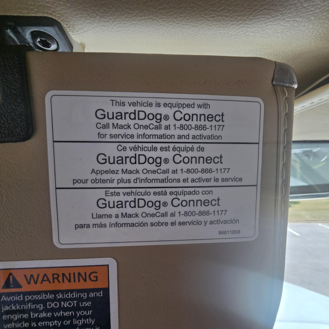Mack PI64 cab interior showing GuardDog Connect sticker with multi-language text and a warning label at bottom left.