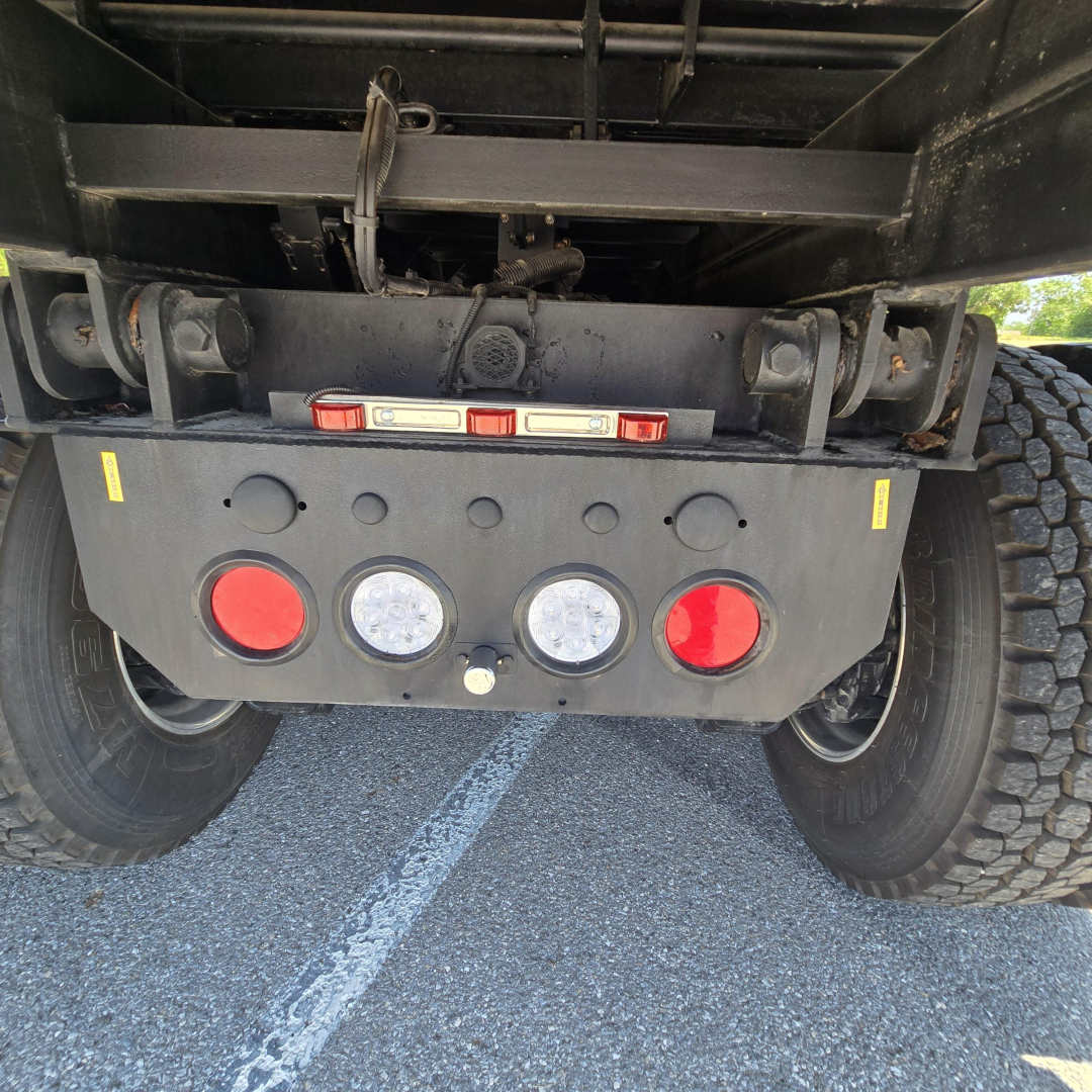 Rear view of a 2026 Mack PI64 dump truck showing bumper with two red reflectors, two large lights, and a level of the hitch area.