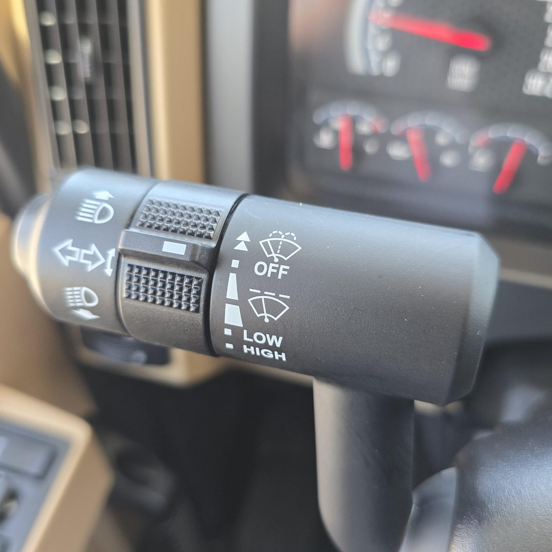 Truck steering column stalk showing headlight and wiper controls in a Mack PI64 cab. Visible text includes OFF, LOW HIGH, and symbols for headlights and wipers.