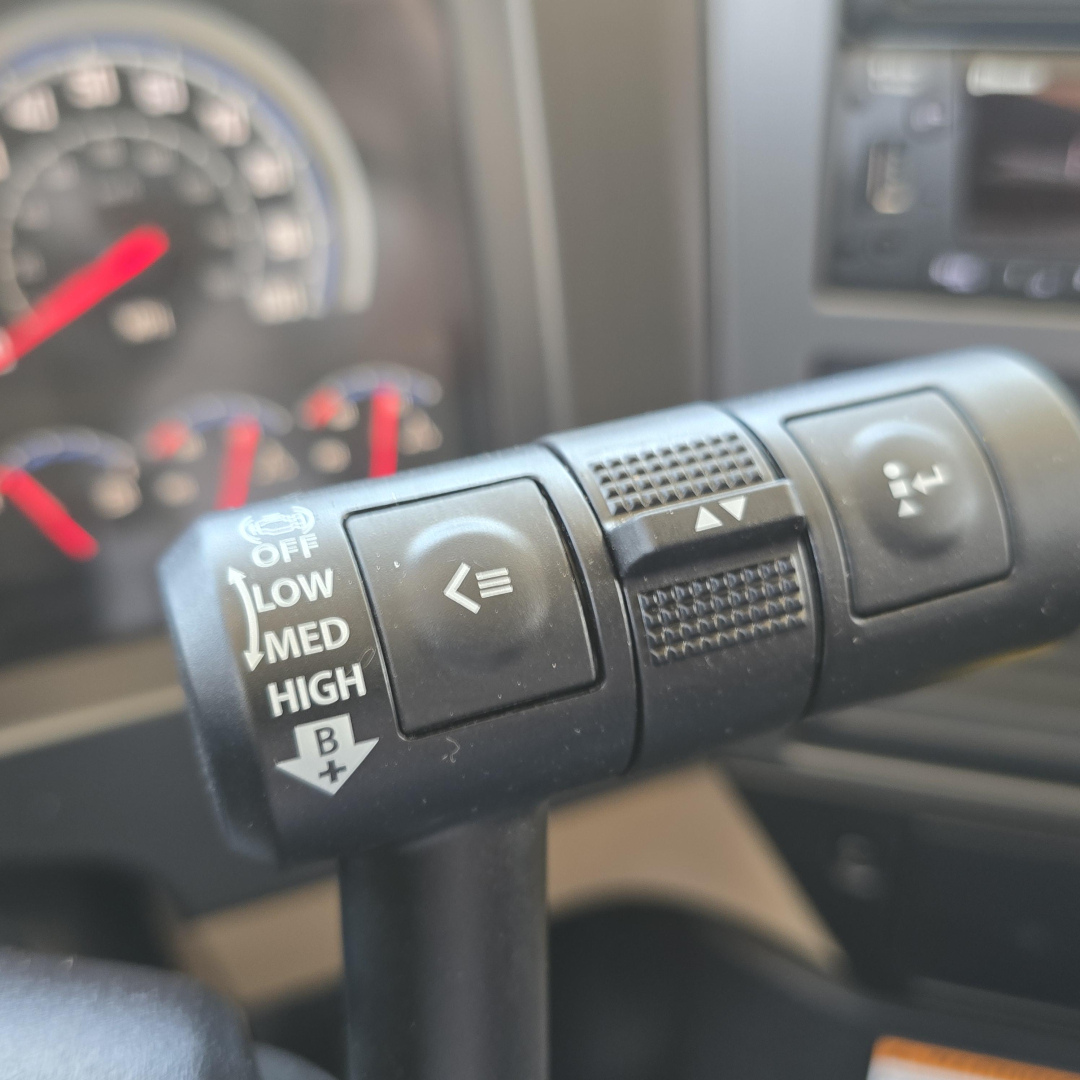Steering column stalks in a Mack PI64 cab showing multi-function controls including light/turn signal lever with ‘OFF LOW MED HIGH’ markings and BL/ arrow, plus adjacent rocker buttons.