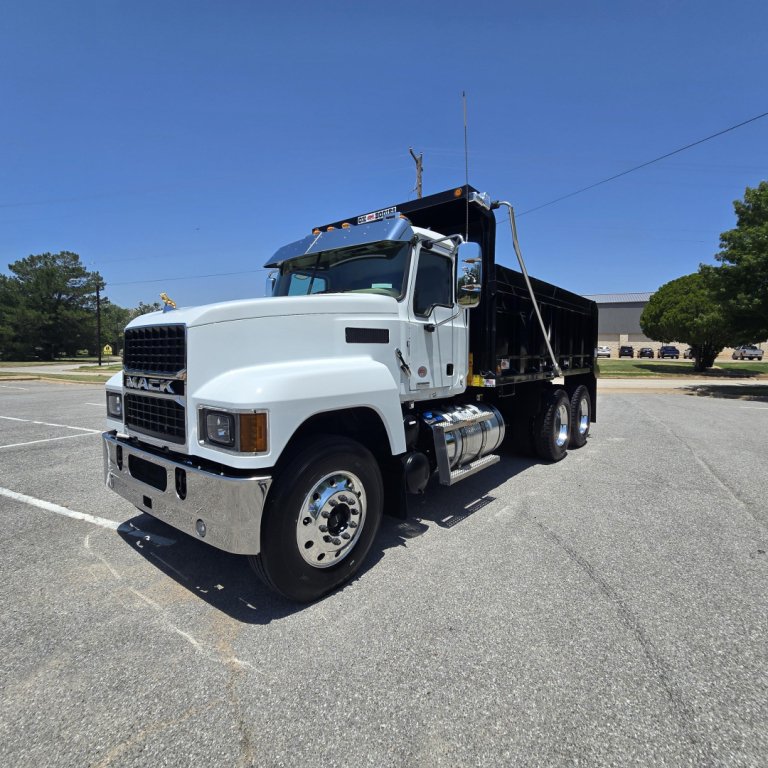 Dump truck shown at angle with white cab and black dump bed, Mack badge on grille, Dual rear wheels, daylight setting.
