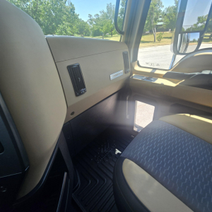 Truck cab interior of a 2026 Mack PI64 showing beige dashboard, vent, and driver's seat with patterned fabric, front window view.