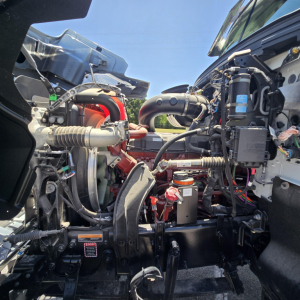 Engine bay of a 2026 Mack PI64 dump truck showing dual air intake hoses, turbocharger, coolant reservoir, wiring harnesses, and various engine components.