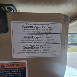 Mack PI64 cab interior showing GuardDog Connect sticker with multi-language text and a warning label at bottom left.
