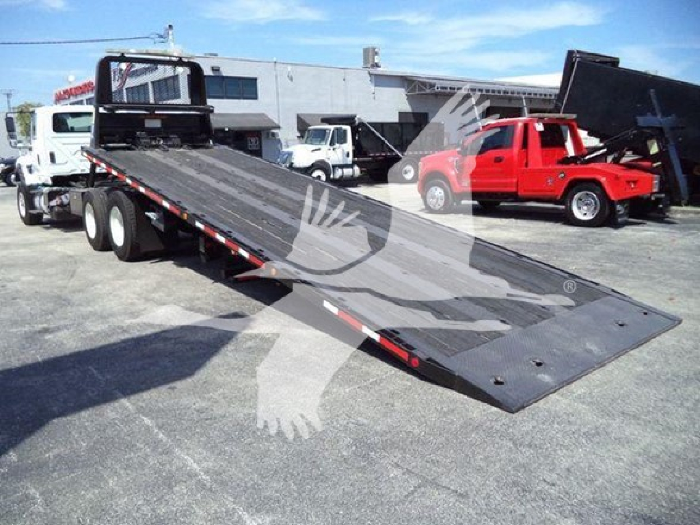 Flatbed tow truck trailer ramp section in a dealership lot with other trucks in background.
