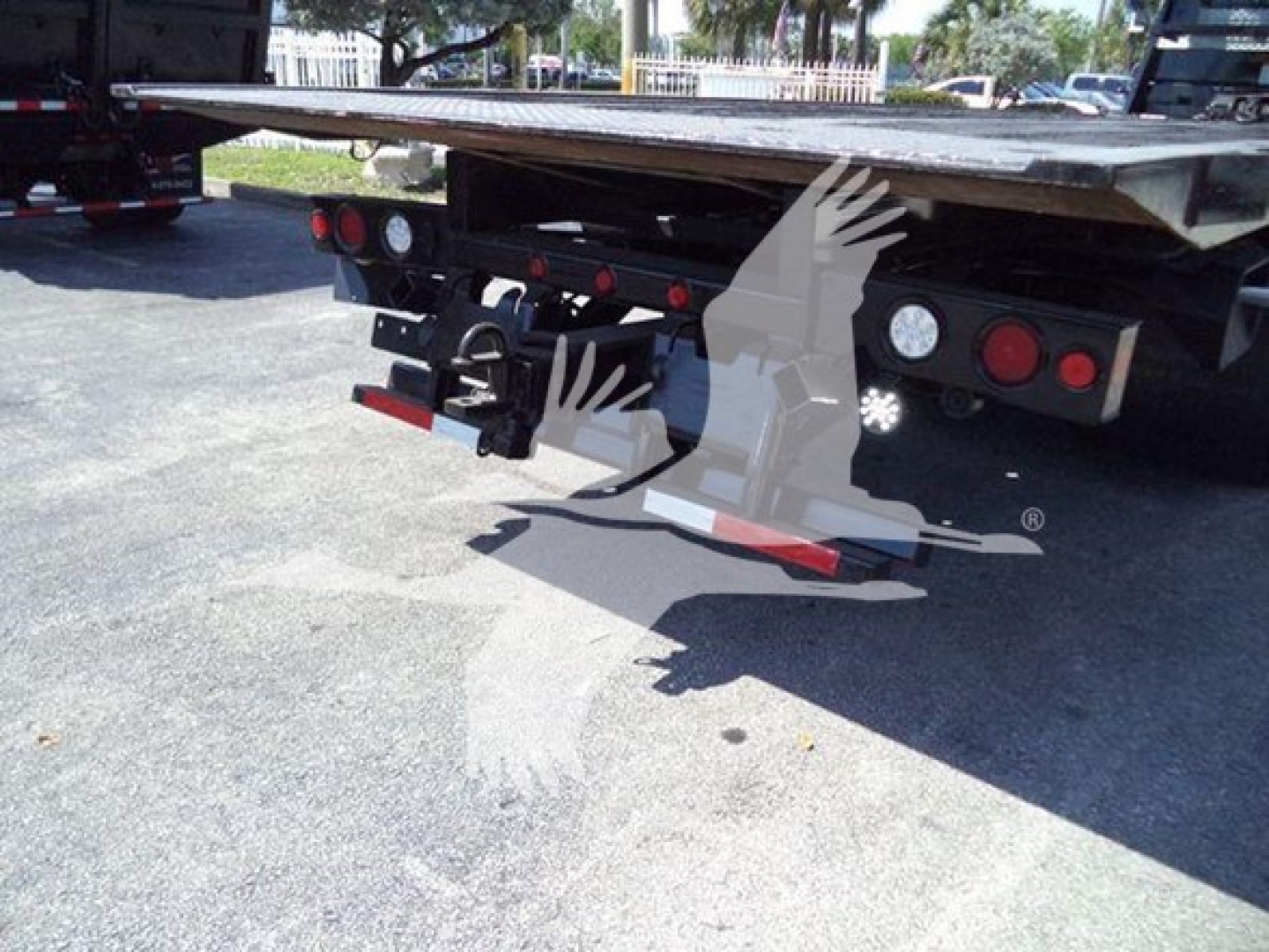 Flatbed tow truck rear with taillights and hitch, platform extended, with red reflectors and bumper; visible metal frame and wheels in background.
