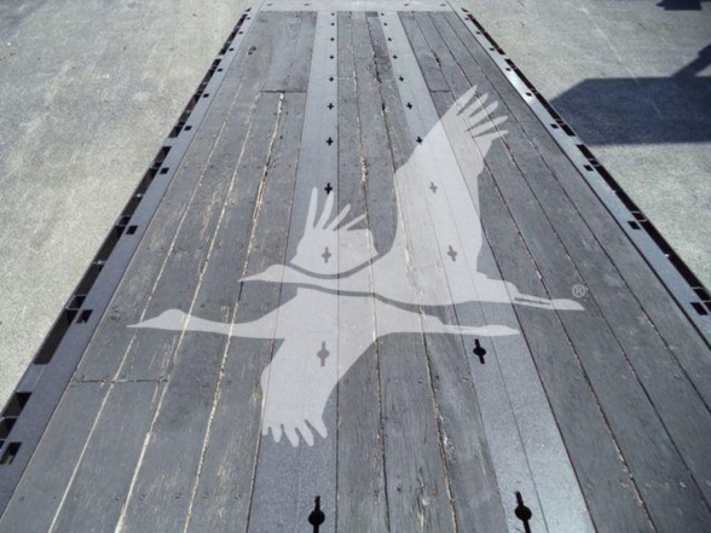 Tractor-trailer flatbed area of a rollback truck showing a wooden deck with metal strips and a watermark of two wings in the center.