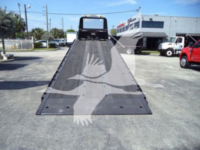 Flatbed tow truck ramp in view from rear, showing long metal deck with hinge at front and black surface, surrounding lot with buildings and other trucks.