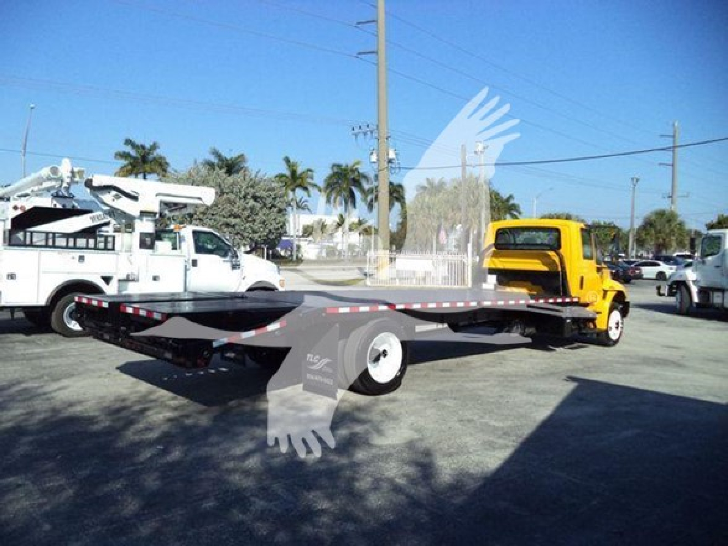 Flatbed tow truck with a yellow cab, long black flatbed, and white utility vehicle in background. No legible text readable on equipment.