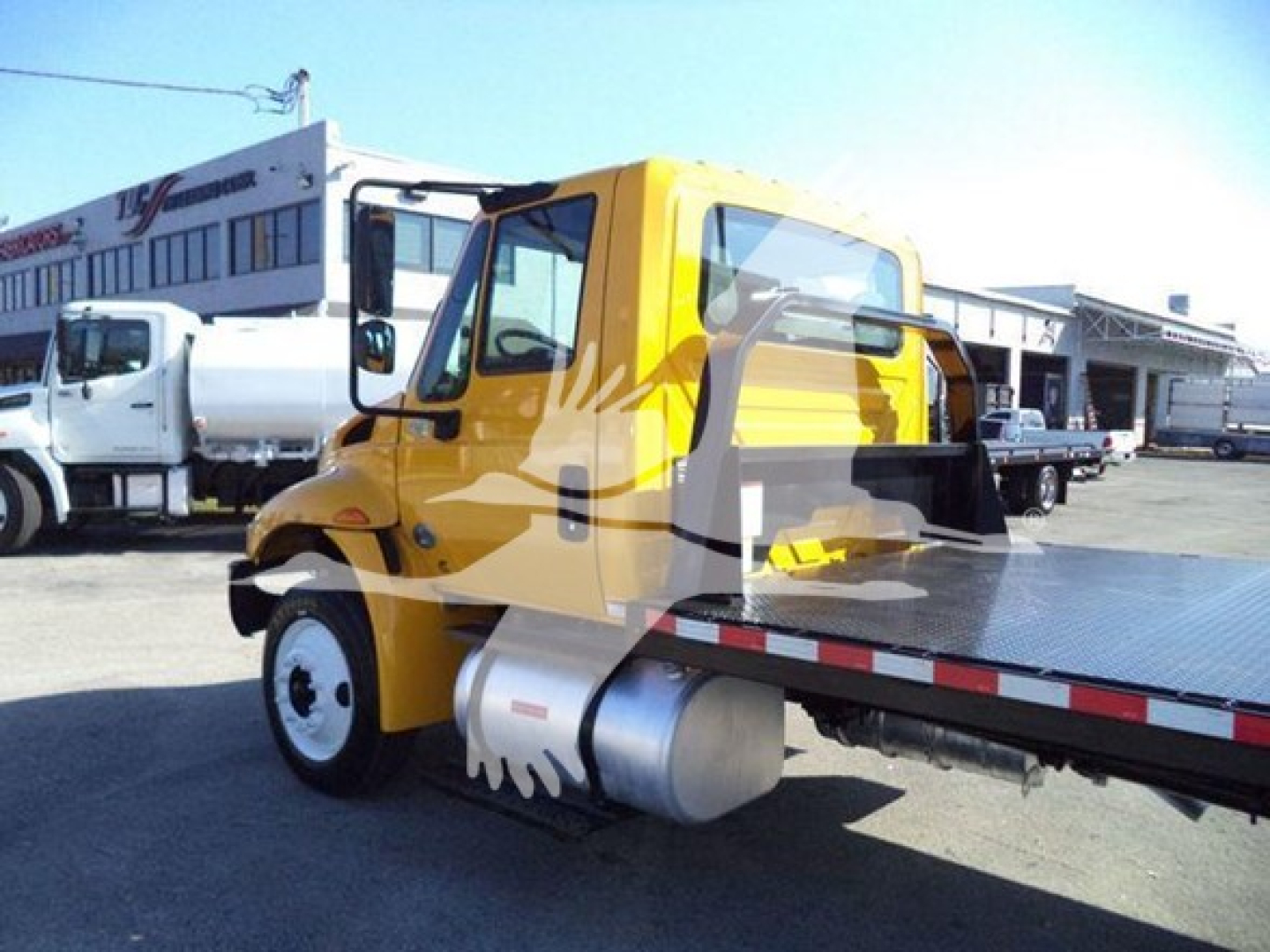 Flatbed tow truck style International 4300 cab in yellow with chrome fuel tank, viewed from rear-left; other trucks in lot in background.