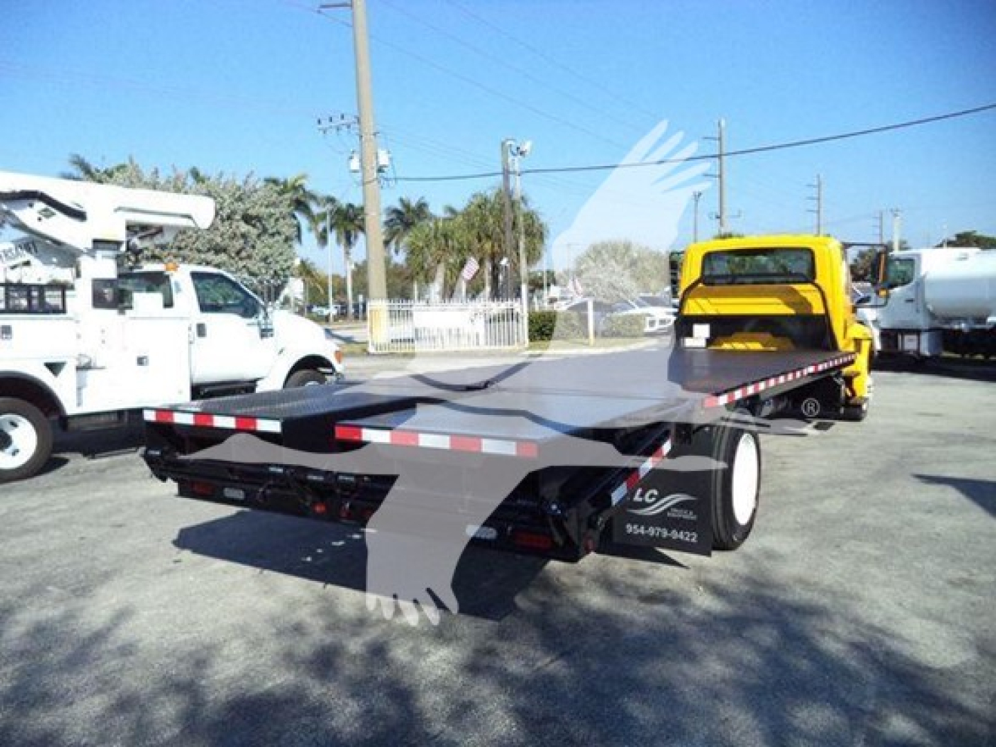 Flatbed tow truck trailer in an industrial lot with a yellow vehicle in background; visible side decals and wheel, no license plate details readable.