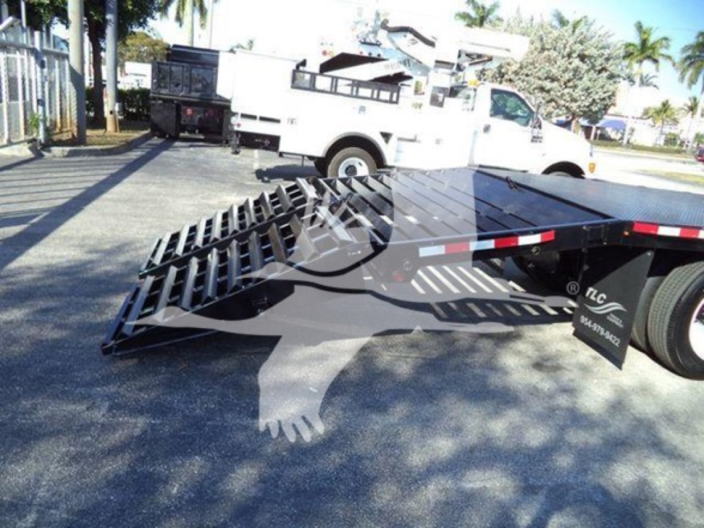 Flatbed tow truck trailer with lowered ramp in foreground, International 4300 service truck in background, equipment yard scene.