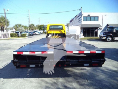 Truck bed platform of a 2019 International 4300 auto carrier seen from rear; yellow tow vehicle and dealership building in background; black flatbed with reflective red/white markings.