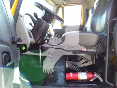 Cab interior of a 2019 International 4300 auto carrier showing steering wheel, driver seat, pedals, and a red fire extinguisher mounted under the dashboard.