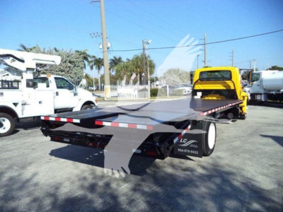 Flatbed tow truck trailer in an industrial lot with a yellow vehicle in background; visible side decals and wheel, no license plate details readable.