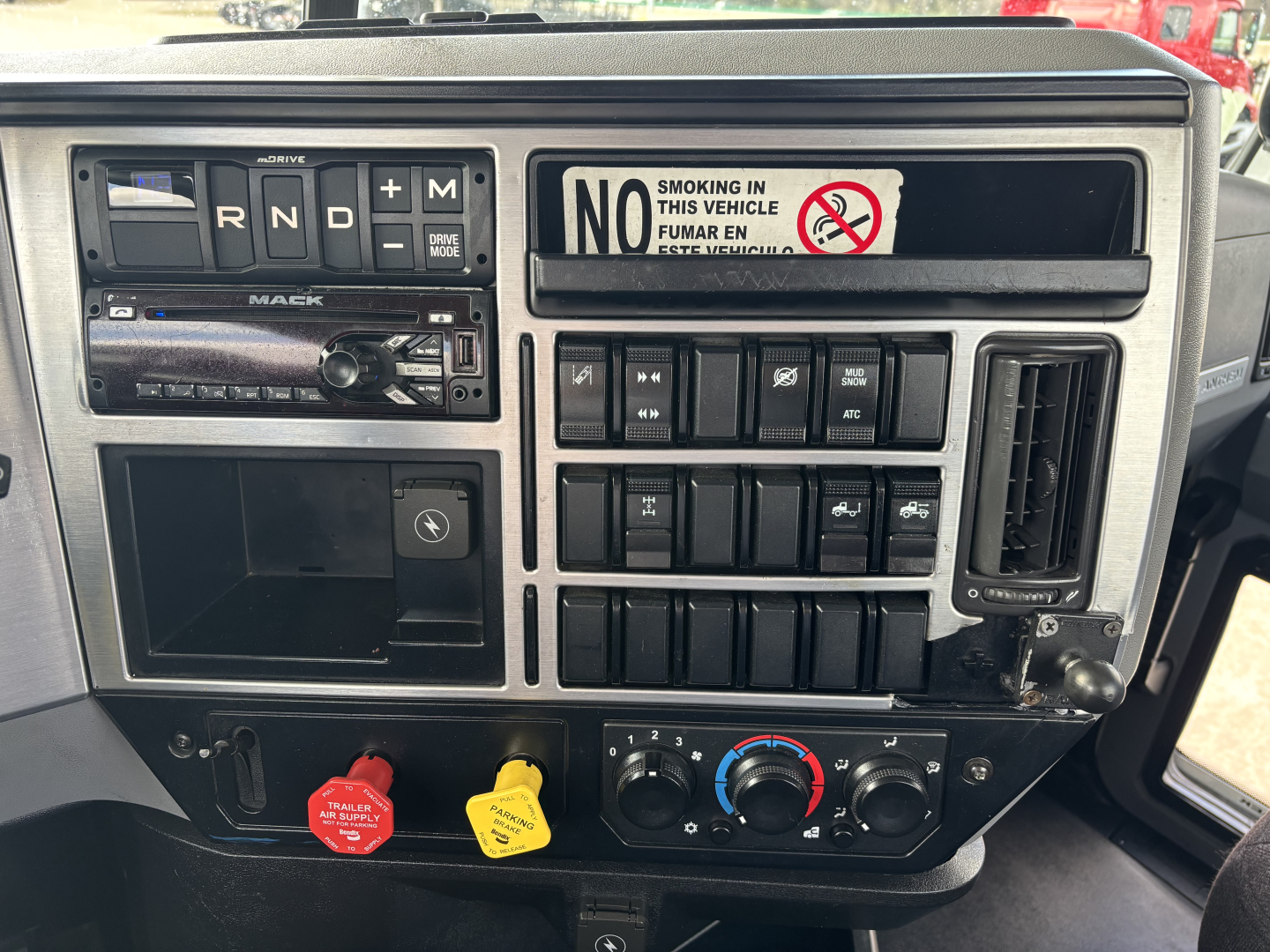 Mack AN64T truck dashboard showing steering column buttons, gear selector with N R D M positions, dashboard switches, climate controls, orange and yellow warning knobs, radio, and a no-smoking sticker.