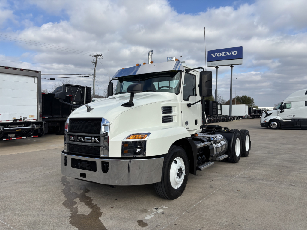 White Mack AN64T conventional daycab truck on lot with diesel exhaust and Volvo sign in background. Front grille shows MACK letters, bumper chrome, dual rear axles.
