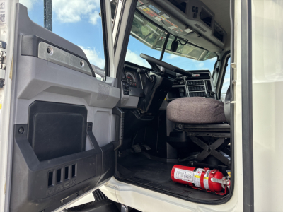 Truck cab interior photo showing driver seat, dashboard, steering wheel, and a fire extinguisher on the floor; open door with steps and window.
