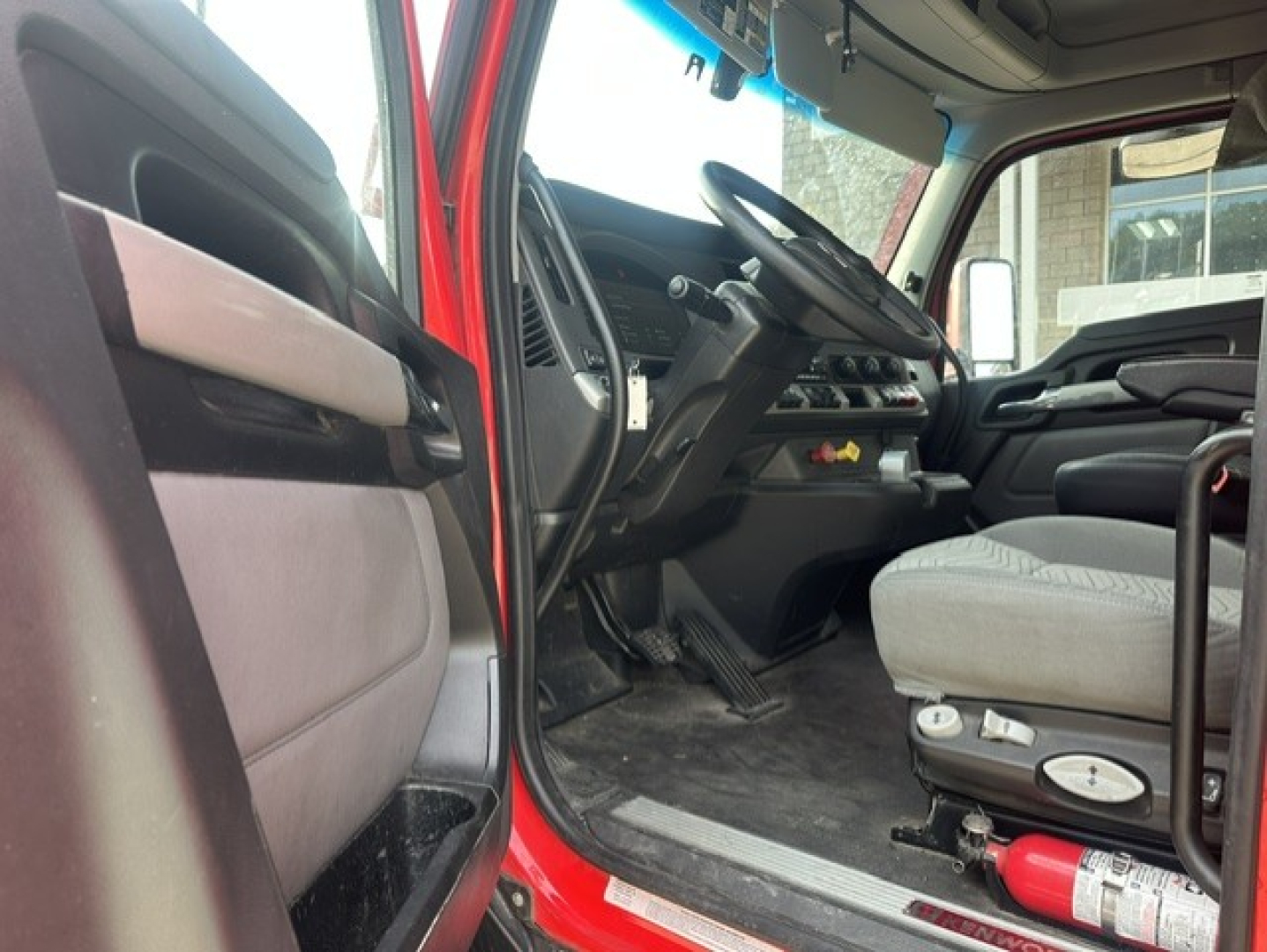 Kenworth T680 truck interior showing steering wheel, driver seat, and dashboard with pedal area; red exterior visible at door frame.