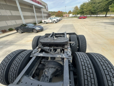 Kenworth T680 chassis cab view showing rear tandem axles and drivetrain, with truck lot and building in background.