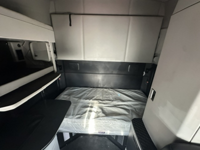 Seat area of a 2023 Kenworth T680 sleeper cab showing a single sleeper bed with plastic covering and storage compartments along the wall.