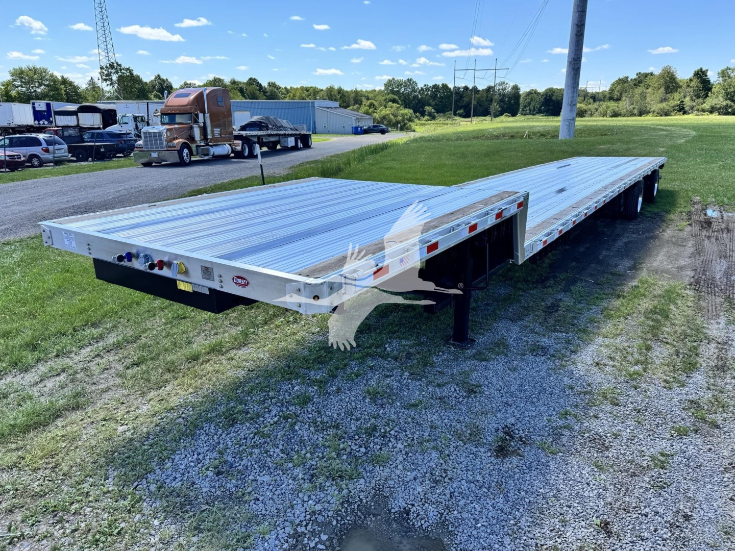 Flatbed trailer showing dual-axle aluminum deck with rear ramp; DOT lights and reflectors along edge; VIN and stock labels on front corner; outdoor lot with other trucks.
